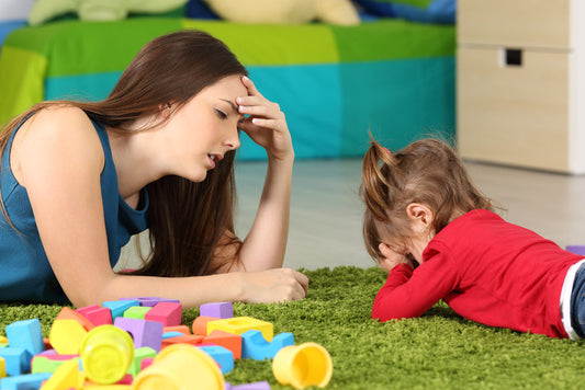 An upset toddler lying on the floor surrounded by toys, with a frustrated-looking woman nearby, illustrating the challenges of taming infant tantrums.